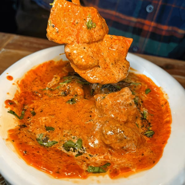 A skewer of chicken tikka being dipped into a bowl of creamy orange curry sauce.