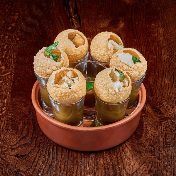 Six pani puri in shot glasses with filling and cilantro, served in a terracotta bowl on wood.