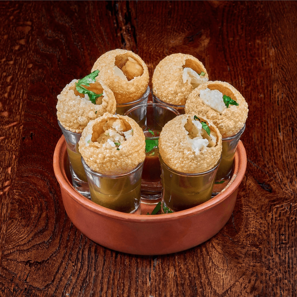 Six pani puri in shot glasses with filling and cilantro, served in a terracotta bowl on wood.